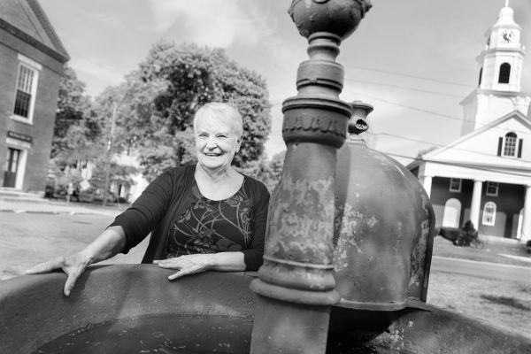 Woman standing behind fountain on Montague Center common with library and church behind her.
