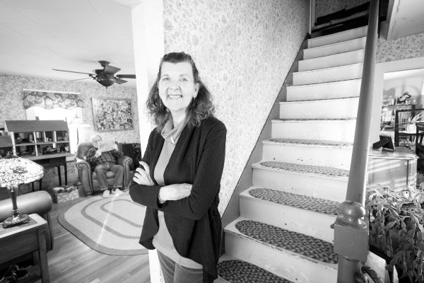 White woman standing with arms crossed in front of stairs with white man reading newspaper in the background. Interior of house.