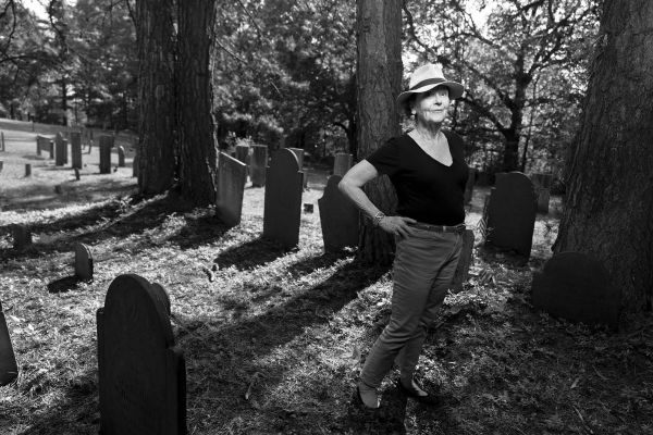 White woman in hat with hands on hips standing in cemetery.