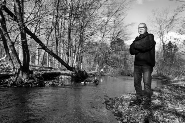 White man with arms crossed standing beside a stream.