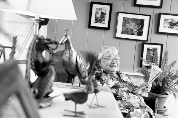Woman with short white hair and glasses reading in front of a wall of framed photographs and a potted plant. In the foreground a table of metal sculptures of animals and a lamp.